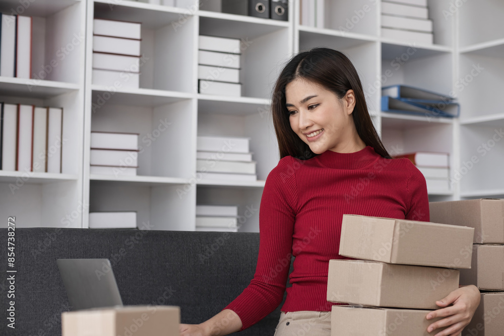 Female Business Owner Managing Online Sales, Packing Products, and Working on Laptop in Modern Office Space with Shelves in Background Female Business Owner Managing Online Sales, Packing Products, and Working on Laptop in Modern Office Space with Shelves in Background