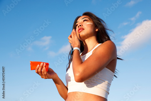 young woman applying sunscreen on her face, skincare in front of blue sky. Product photography concept