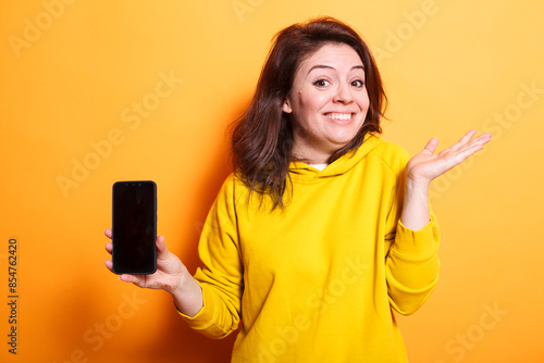 Excited woman wearing casual attire holding a smartphone, grinning broadly, and announces a new device and good news against isolated backdrop. Promotional idea with copyspace. photo