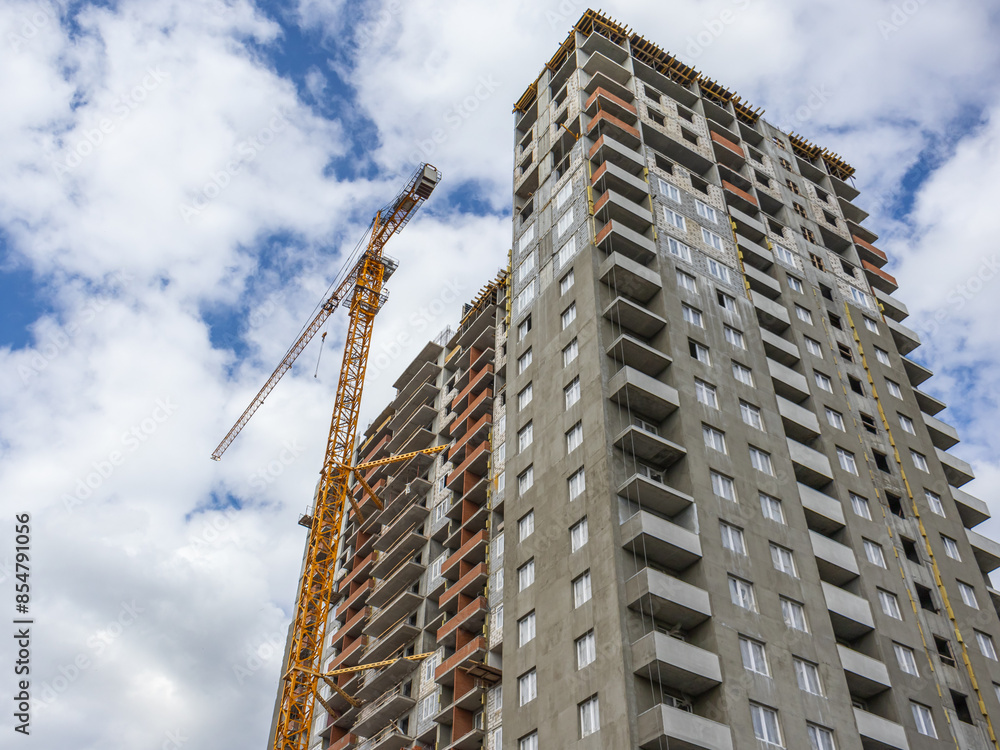 Fototapeta premium A tower construction crane on the background of a blue sky with clouds. The frame of a building under construction. Construction works. The facade of the building is plastered with a cement mixture.