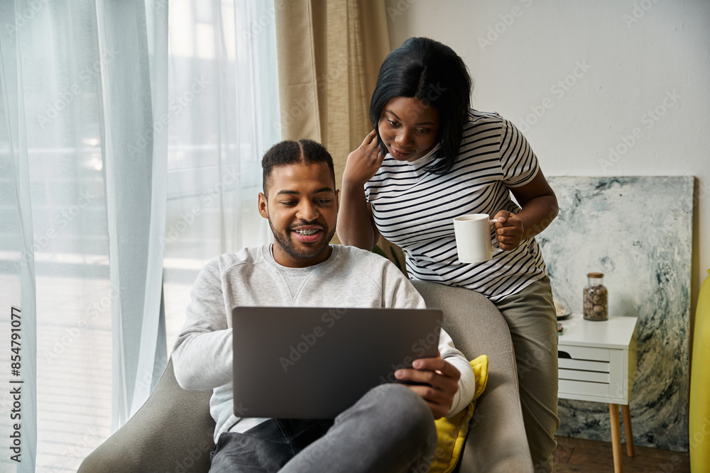 Fototapeta premium African American couple cozy at home, using laptop and enjoying drink.