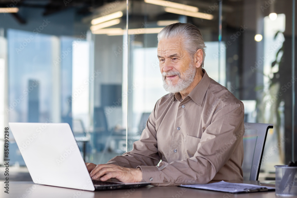 Senior man working on a laptop in a modern office, demonstrating focus and productivity. Professional work environment with glass walls and natural light. Senior man working on a laptop in a modern office, demonstrating focus and productivity. Professional work environment with glass walls and natural light.