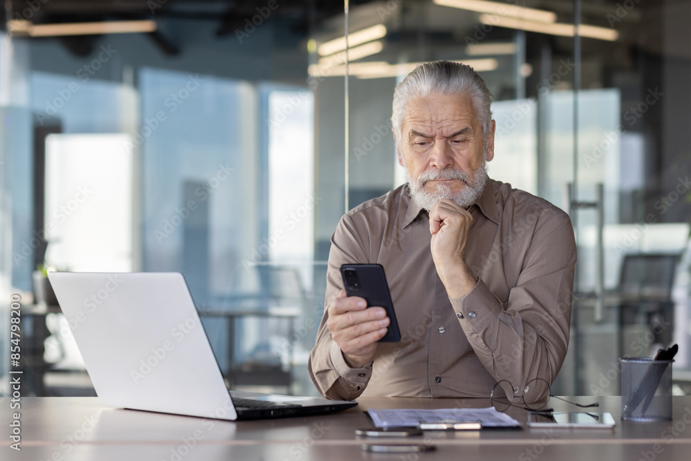 Elderly businessman working in a modern office, using a smartphone to analyze data, with a laptop in front of him Elderly businessman working in a modern office, using a smartphone to analyze data, with a laptop in front of him