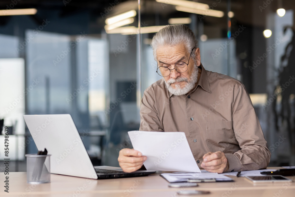 Senior businessman reviewing documents while sitting at a desk with a laptop in a modern office setting. Senior businessman reviewing documents while sitting at a desk with a laptop in a modern office setting.
