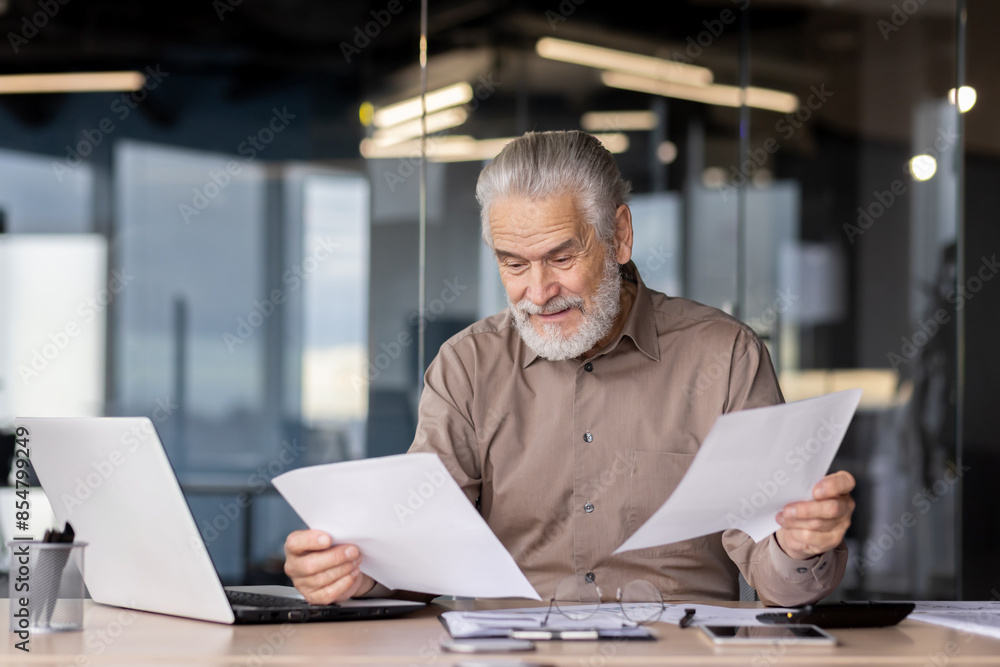 Senior businessman in a modern office reviewing paperwork at his desk with a laptop and reading glasses Senior businessman in a modern office reviewing paperwork at his desk with a laptop and reading glasses