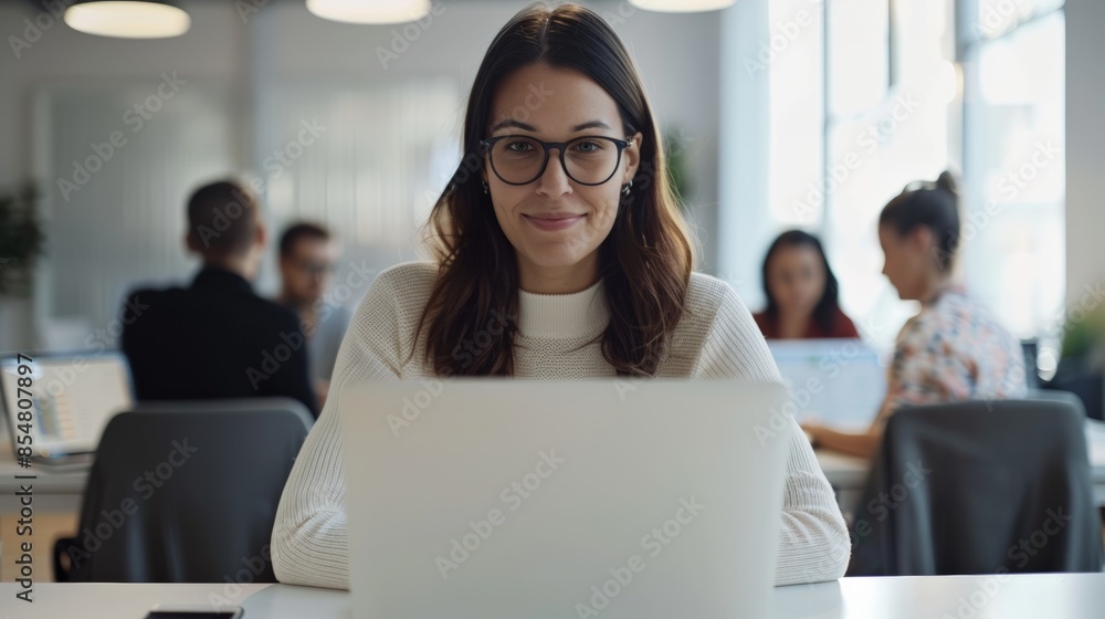 Young professional woman working on laptop in modern office setting with colleagues in the background, focused and productive Young professional woman working on laptop in modern office setting with colleagues in the background, focused and productive