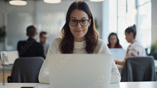 Young professional woman working on laptop in modern office setting with colleagues in the background, focused and productive