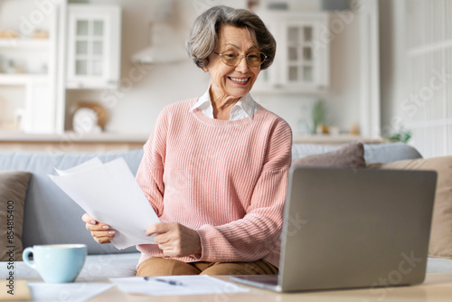 Positive elderly woman, sitting on sofa with paper receipt in her hands, calculating expenses, managing family budget with laptop