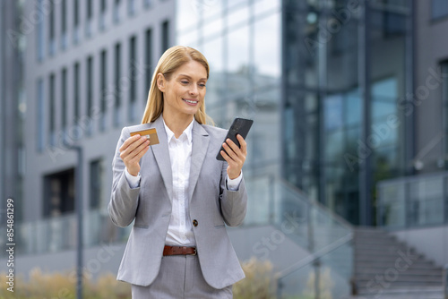 Smiling young businesswoman in suit standing outside, holding golden credit card and using mobile phone smiling.