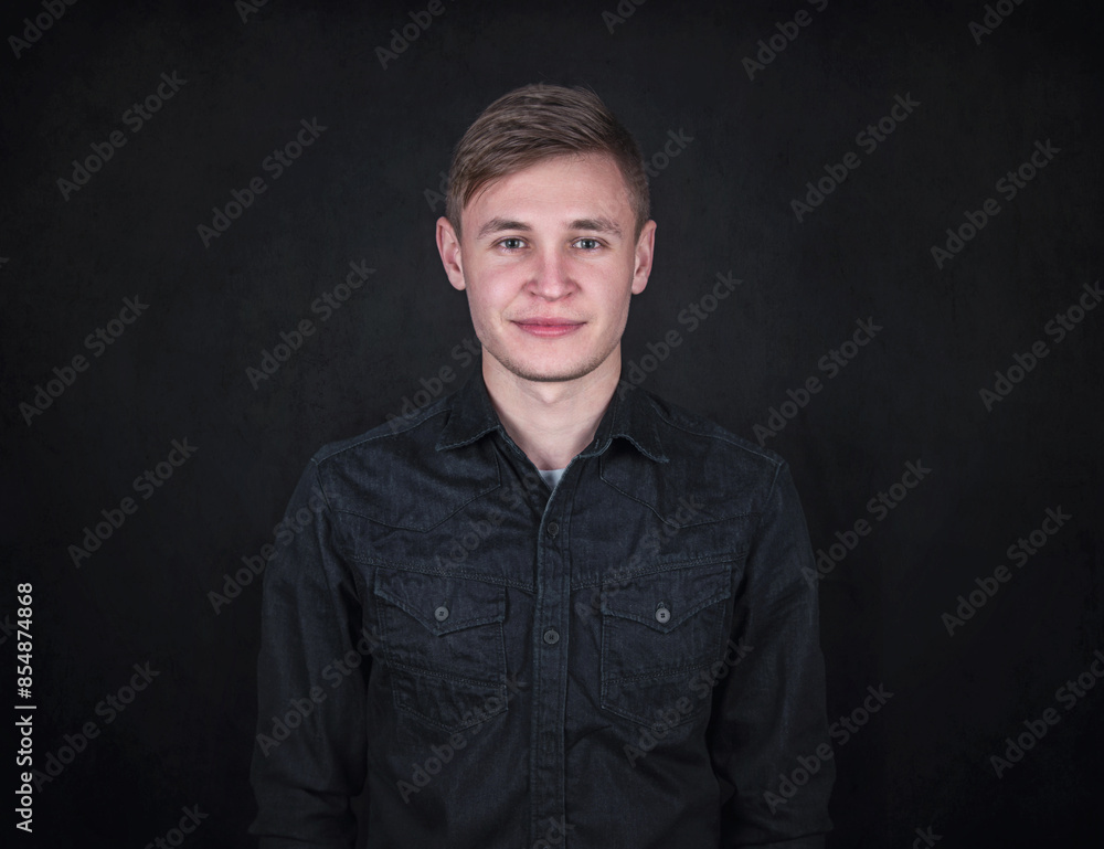 Portrait of a carefree young man against a black wall background looking to camera. Relaxed male model with joyful face expression wearing dark blue jeans jacket Portrait of a carefree young man against a black wall background looking to camera. Relaxed male model with joyful face expression wearing dark blue jeans jacket
