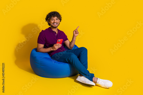 Full body photo of young man sit bean bag hold device point empty space dressed stylish violet clothes isolated on yellow color background photo