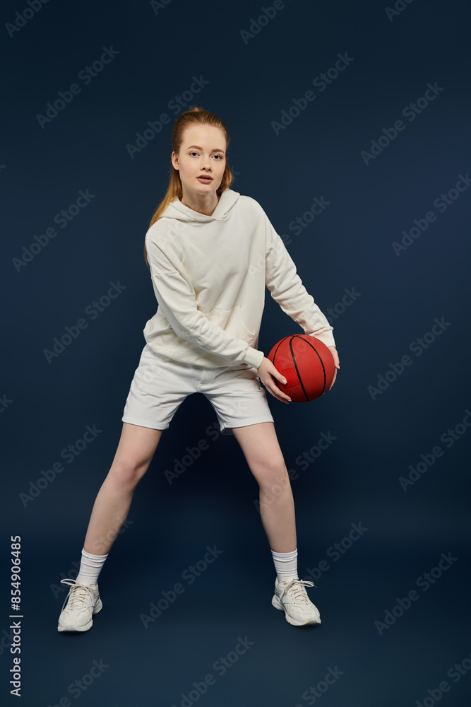 A young woman in a white hoodie and shorts holds a basketball in front of a blue backdrop. A young woman in a white hoodie and shorts holds a basketball in front of a blue backdrop.