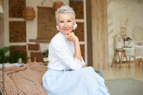 Stylish mature female in elegant fashionable clothes posing of boho style living-room, dressed in long skirt, hand-made laced blouse and flower-shaped jewelry in ears. Female fashion, interior design