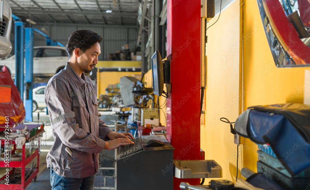 Young mechanic in work uniform is using a computer in an auto repair shop with tools and vehicle in the background. Young mechanic in work uniform is using a computer in an auto repair shop with tools and vehicle in the background.