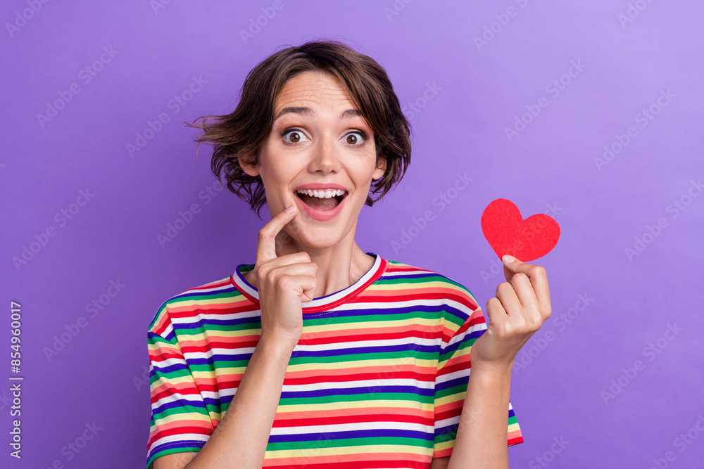 Portrait of cute astonished person finger touch chin arm hold little red paper heart isolated on purple color background Portrait of cute astonished person finger touch chin arm hold little red paper heart isolated on purple color background