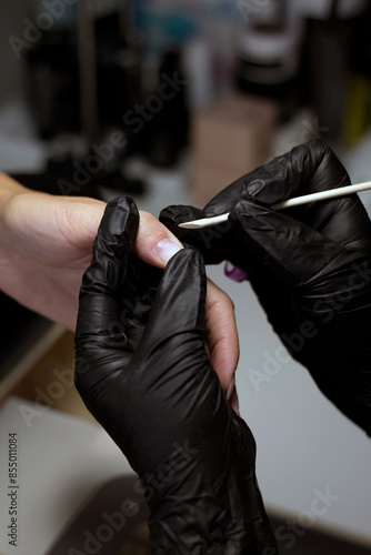 A detailed view of a manicurist’s hands in black gloves, using a wooden stick to clean a client’s nails, emphasizing the precision and care in nail treatment. © Liana