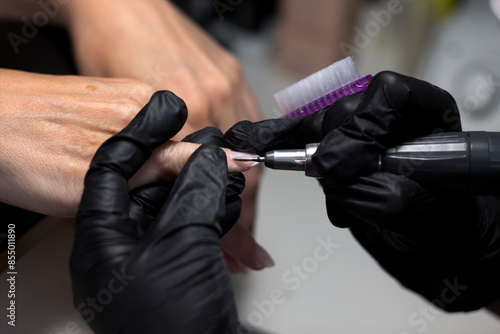 A manicurist in black gloves carefully uses an electric file on a client’s nails, showcasing the precision and expertise involved in professional nail grooming. © Liana