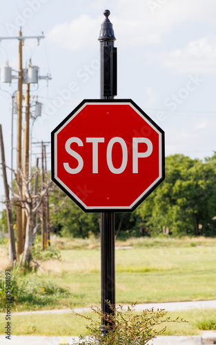A close-up of a stop sign on a black post situated at the edge of a grassy area with an urban background. 