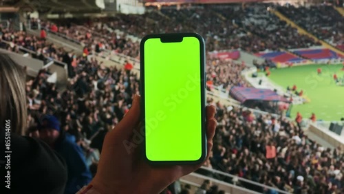 A woman watches football at the stadium, holds a phone with a green screen in hands. Makes bets on sports.