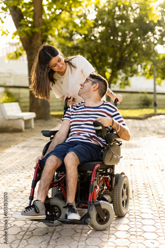 A young woman cares for her disabled brother in an automatic wheelchair at an outdoor park. Disabled woman and man look at each other happily.Concept of men with cerebral palsy.