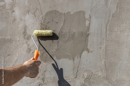 A worker applies the primer to the wall with a long-handled roller. A man's hand rolls a primer onto a gray wall. Wet footprint on a plastered wall. House facade renovation concept.