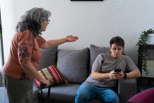 A mother stands and gestures emphatically while her adult son, sitting on the couch, looks at his phone, ignoring her, highlighting a moment of family tension and communication breakdown. photo