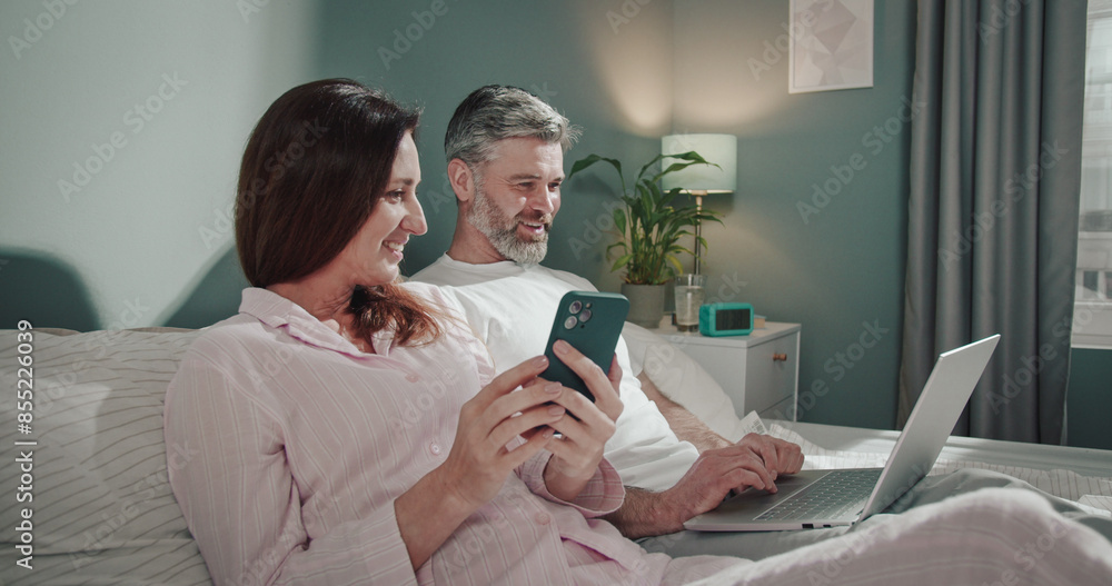 Beautiful happy caucasian adult couple relaxing on bed at home, man working on laptop computer while his cute wife reading something at the smartphone. People and gadgets concept Beautiful happy caucasian adult couple relaxing on bed at home, man working on laptop computer while his cute wife reading something at the smartphone. People and gadgets concept