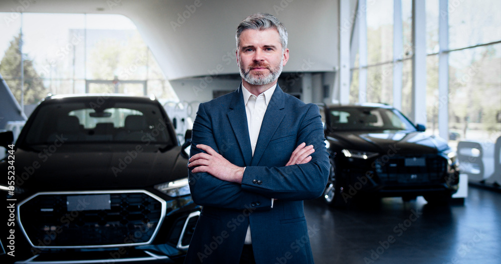 Confident successful car dealer at the background of new automobile in showroom. Handsome bearded caucasian man standing in car dealership, looking at camera and crossing hands. Confident successful car dealer at the background of new automobile in showroom. Handsome bearded caucasian man standing in car dealership, looking at camera and crossing hands.