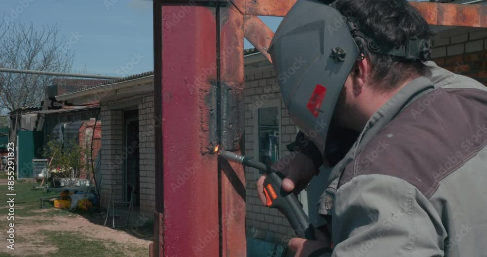 A man in a protective mask repairs an old gate using welding. Having welded the metal, the man evaluates the result. The man takes off his mask and evaluates the quality of the weld. A man in a protective mask repairs an old gate using welding. Having welded the metal, the man evaluates the result. The man takes off his mask and evaluates the quality of the weld.
