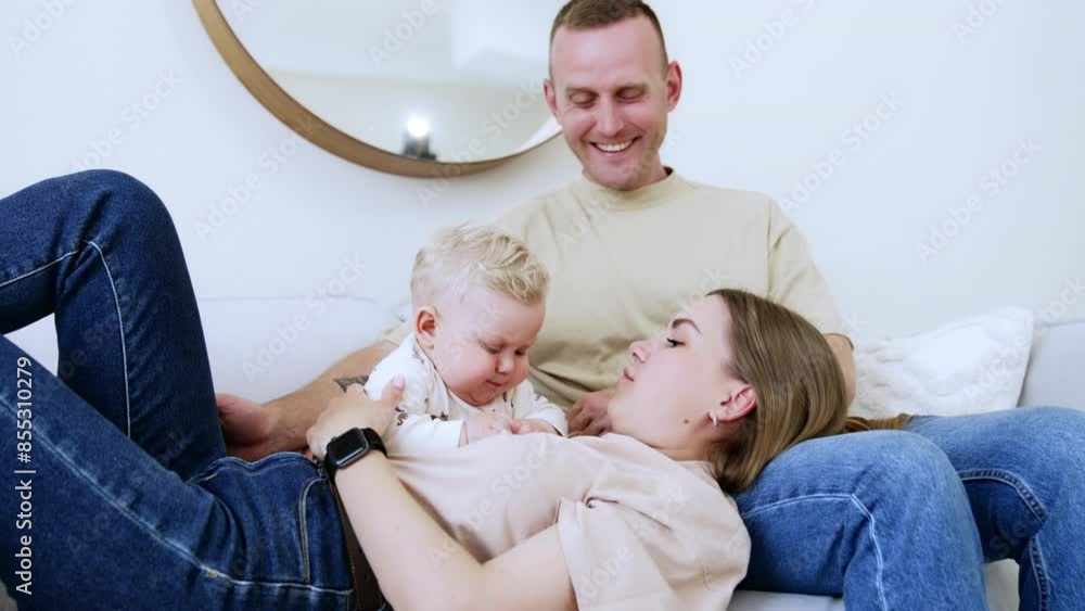 Happy Caucasian parents having rest together with their baby. Little kid sits by his mom lying on husband's laps. Proud dad caresses son's head. Happy Caucasian parents having rest together with their baby. Little kid sits by his mom lying on husband's laps. Proud dad caresses son's head.