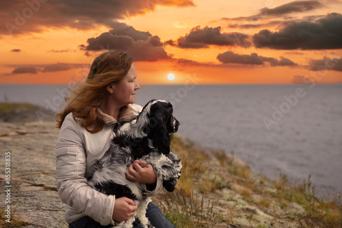 Portrait of a woman and an English Cocker Spaniel puppy.She sits on the rocky shore of the island and holds a dog in her hands.Both look into the distance.In the background there is a lake and sunset. photo