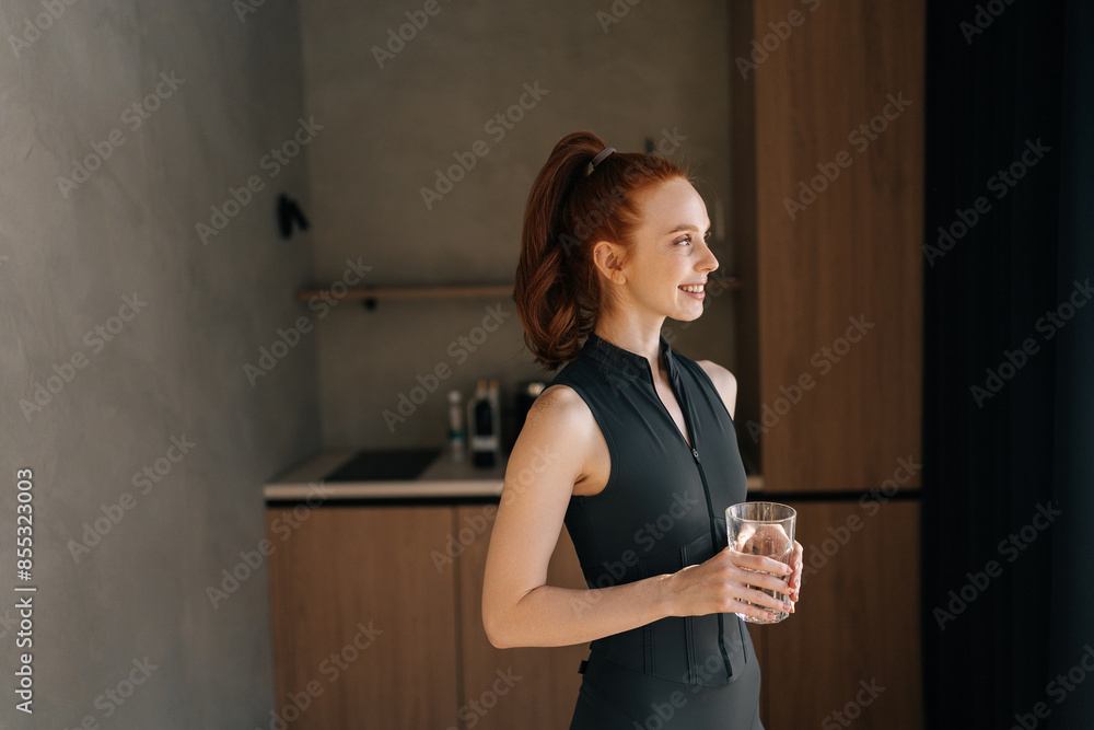 Portrait of positive young sportswoman drinking clean water in cup, satisfied after home training standing by window on sunny morning, smiling looking away. Healthy lifestyle, fitness concept. Portrait of positive young sportswoman drinking clean water in cup, satisfied after home training standing by window on sunny morning, smiling looking away. Healthy lifestyle, fitness concept.