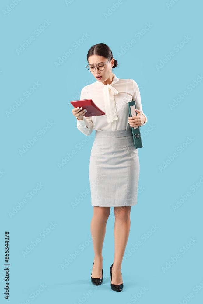 Upset female Asian accountant with calculator and folder on blue background Upset female Asian accountant with calculator and folder on blue background