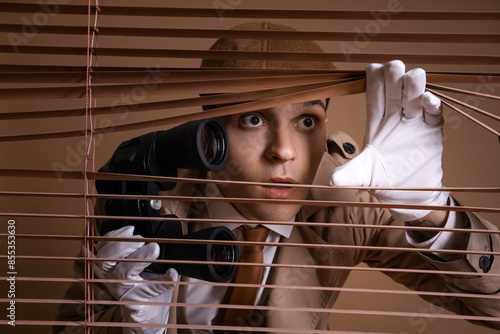 Fotografie Male spy with binoculars looking through blinds on brown background, closeup