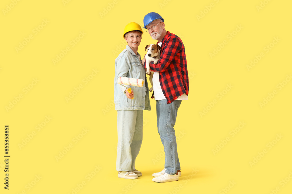 Mature couple in hardhats with dog and paint roller on yellow background Mature couple in hardhats with dog and paint roller on yellow background