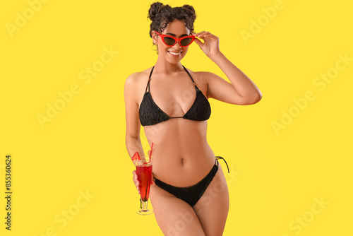 Young African-American woman in swimsuit with cocktail on yellow background