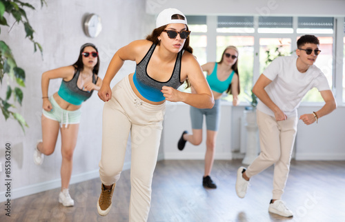 Female teen perform choreographic exercise and teach energetic mobile social dance jazz-funk with friends. Young girls in dark glasses and ball cap guy repeat movements, train in spacious studio