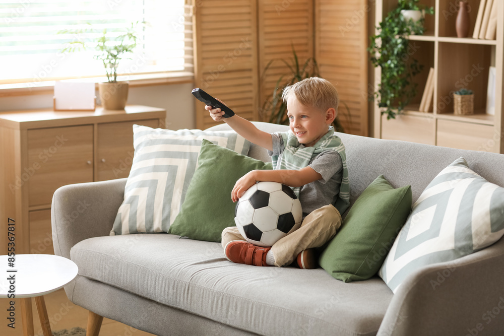 Cute little football player with soccer ball watching TV at home