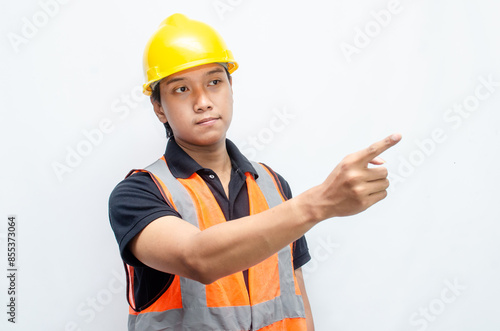 asian man consrtruction worker wearing yellow safety helmet and orange vest smiling and pointing finger to copy space with happy and surprised expression. photo