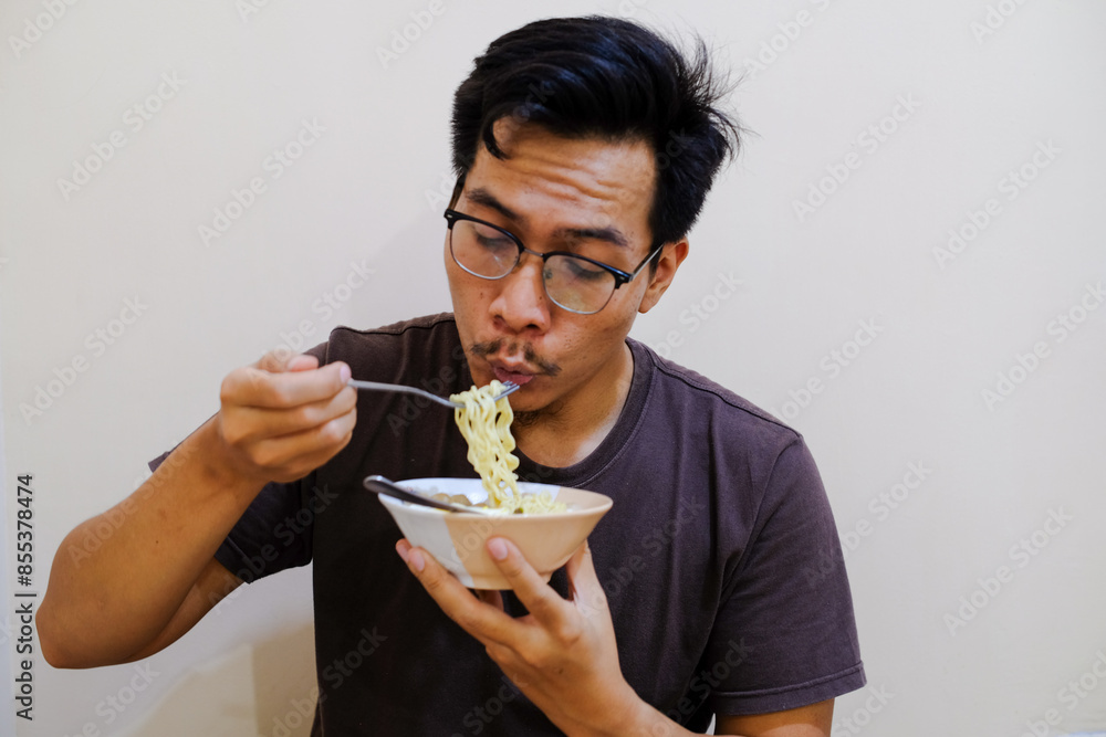 A Southeast Asian man holding and eating noodles in the white bowl. A Southeast Asian man enjoys instan noodles with an expression of satisfaction. A Southeast Asian man holding and eating noodles in the white bowl. A Southeast Asian man enjoys instan noodles with an expression of satisfaction.