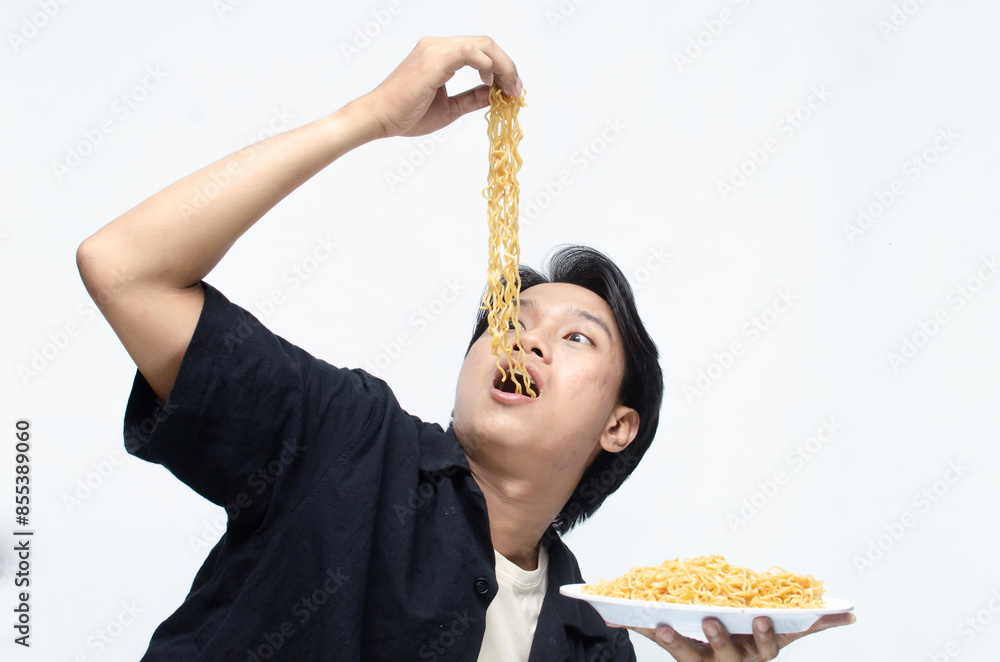 young asian man barehanded pinching fried instant noodles into his mouth. attractive asian man demonstrating how tasty the noodles are. young asian man barehanded pinching fried instant noodles into his mouth. attractive asian man demonstrating how tasty the noodles are.