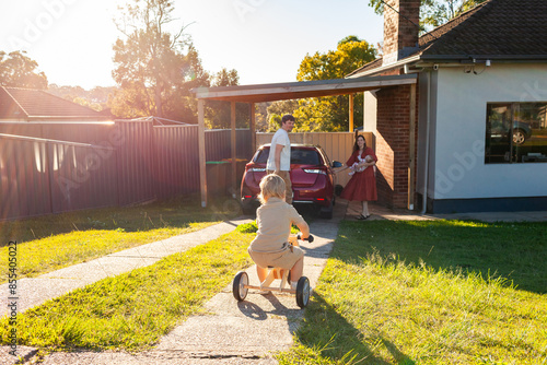 Dad closing car boot while mum holds baby and toddler rides trike on driveway