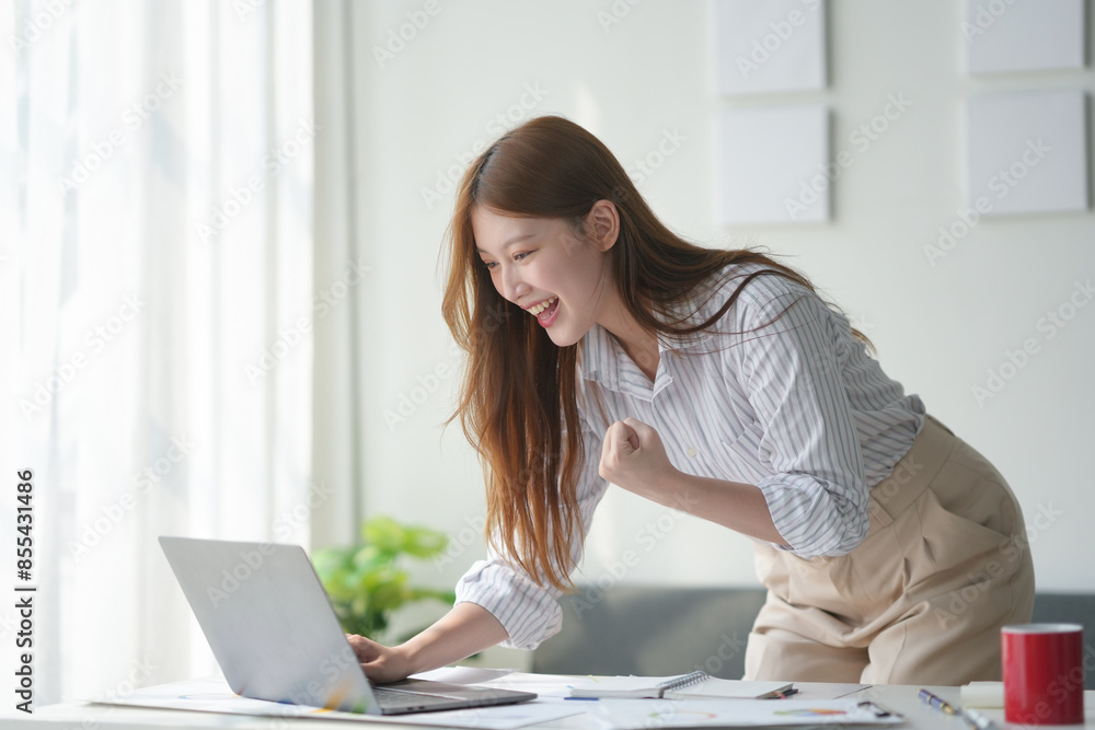Excited businesswoman celebrating success in front of laptop in modern office setting with documents on the desk.