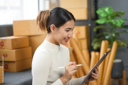 Smiling woman holding a tablet, surrounded by cardboard boxes and shipping supplies, working in a home office. Online business concept. photo