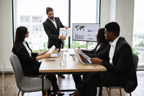 Business team in formal meeting discussing charts and graphs with laptops and documents. Professional presentation in modern office setting with large windows displaying cityscape view.