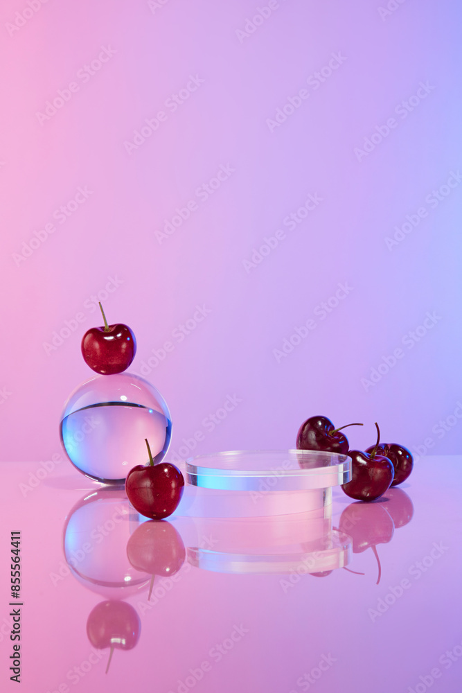 Fototapeta premium The blank showcase pedestal of glass placed in the middle of table, decorated beside with flat lay cherries and a crystal ball, against colorful background. Space for presentation and adding text