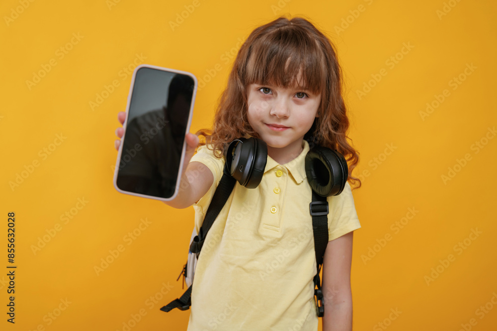 Holding smartphone, headphones on the neck. Cute little girl is against yellow background Holding smartphone, headphones on the neck. Cute little girl is against yellow background
