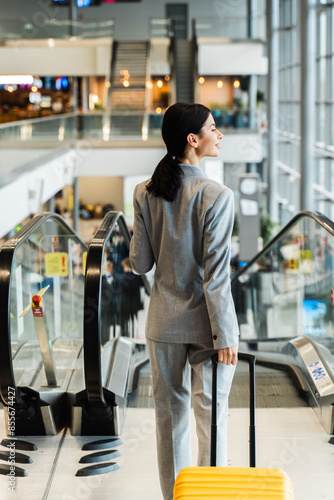 Asian woman with yellow suitcase walks to the escalator in the airport terminal. Journey and vacation concept. Travel blogger is waiting for a new trip in the summer season of 2024.