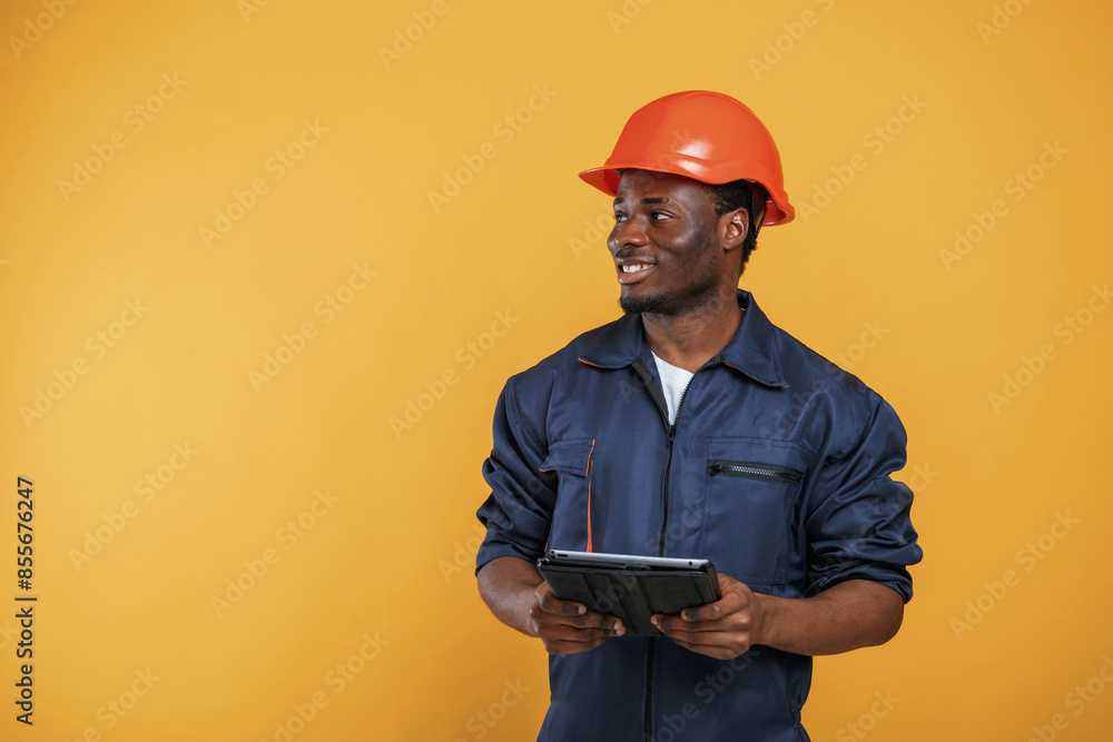 Construction worker in hard hat and with tablet. Handsome black man is in the studio against yellow background Construction worker in hard hat and with tablet. Handsome black man is in the studio against yellow background