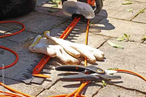 Electric hedge trimmer, pruners and gloves on stone tile in backyard. Garden and hedge maintenance. Nature and equipment. 